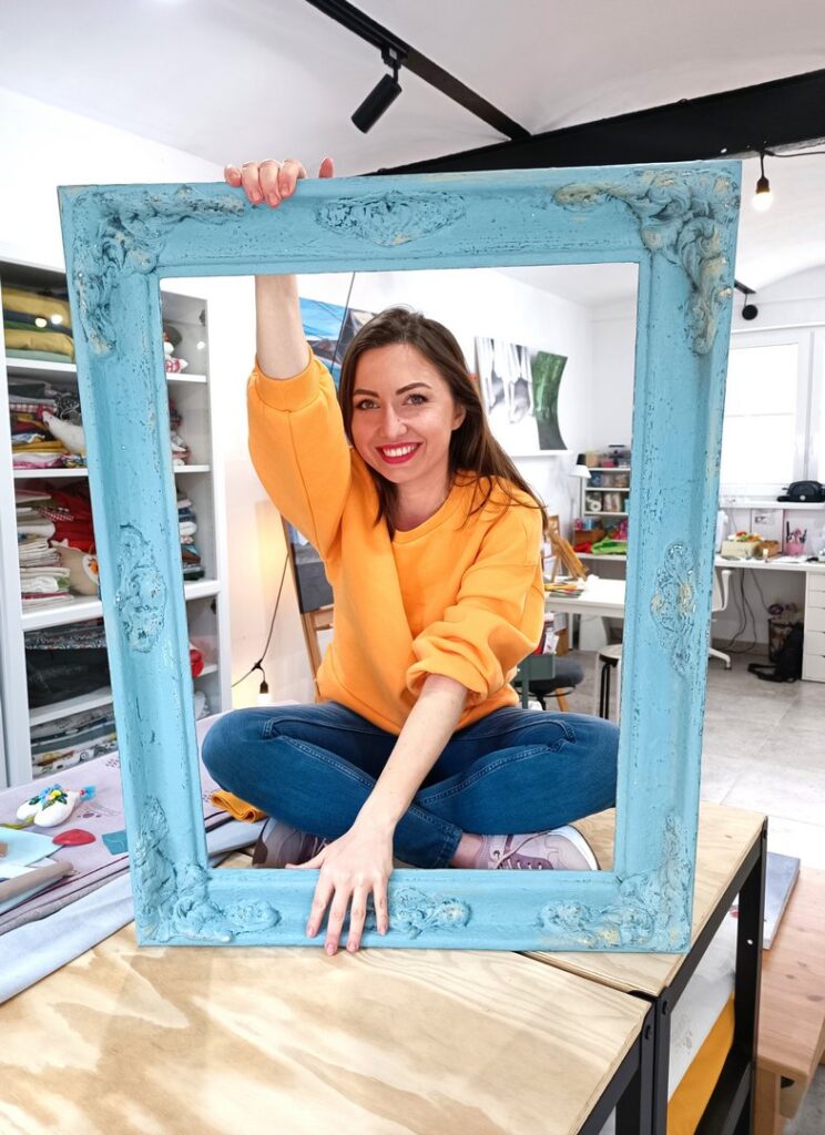 Kasia siedzi na stole w swojej pracowni, trzyma ramę obrazu
